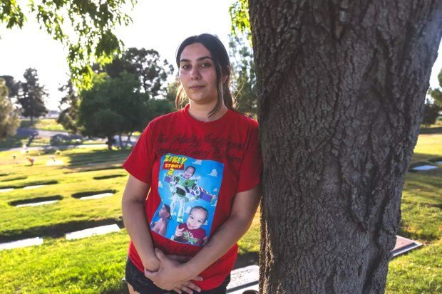 Fentanil krizinde bebekler ve yürümeye başlayan çocuklar şüphesiz kurbanlar haline geliyor 3 A woman stands against a tree in a T-shirt with a boy's face on it